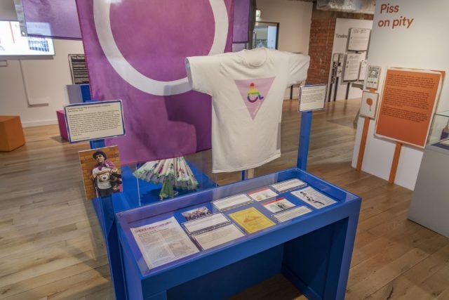 A museum exhibition showing Anis Aktar's Disabled Intersex flag and a white t-shirt with an inverted pink triangle on it and a rainbow-coloured wheelchair user icon.